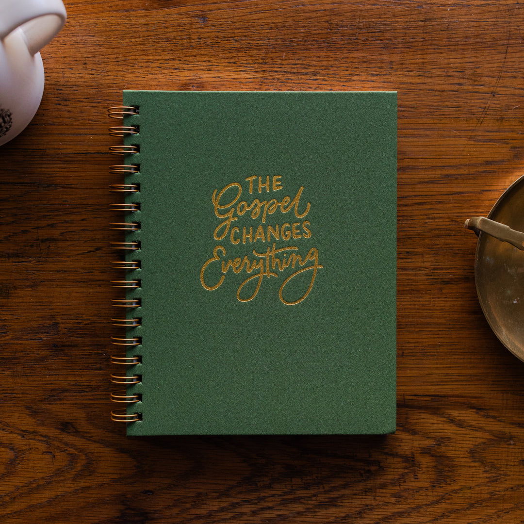 The Gospel Changes Everything Not taking Journal on a wooden table next to a coffee cup and an bronze oil lamp