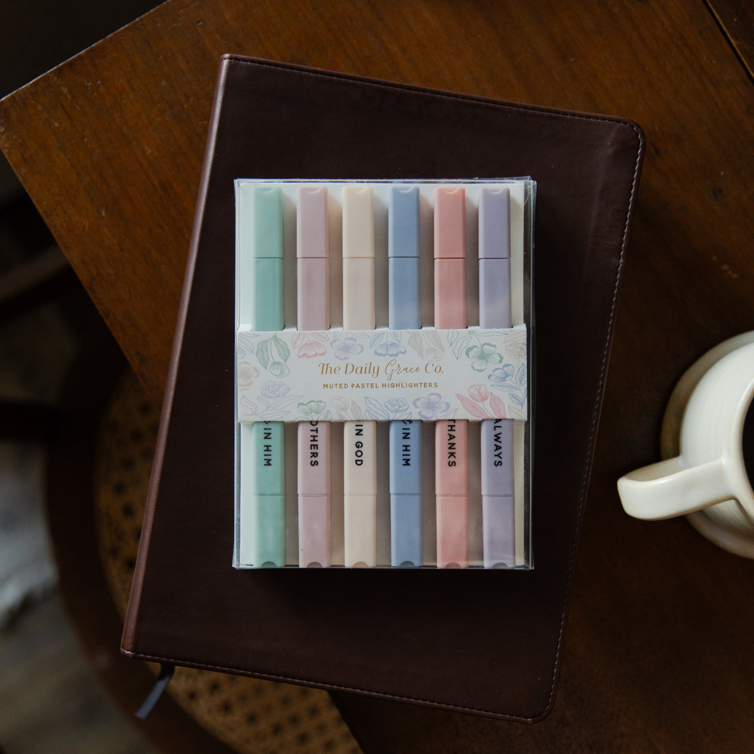 Set of pastel-colored markers in a clear plastic case on a wooden surface with a notebook and mug.