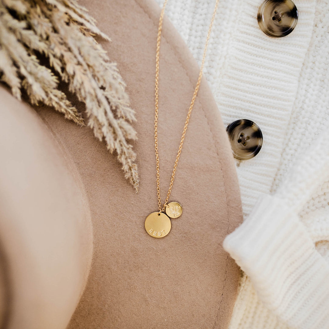 Gold necklace with two pendants that have the words "rest" and the image of a flower. 