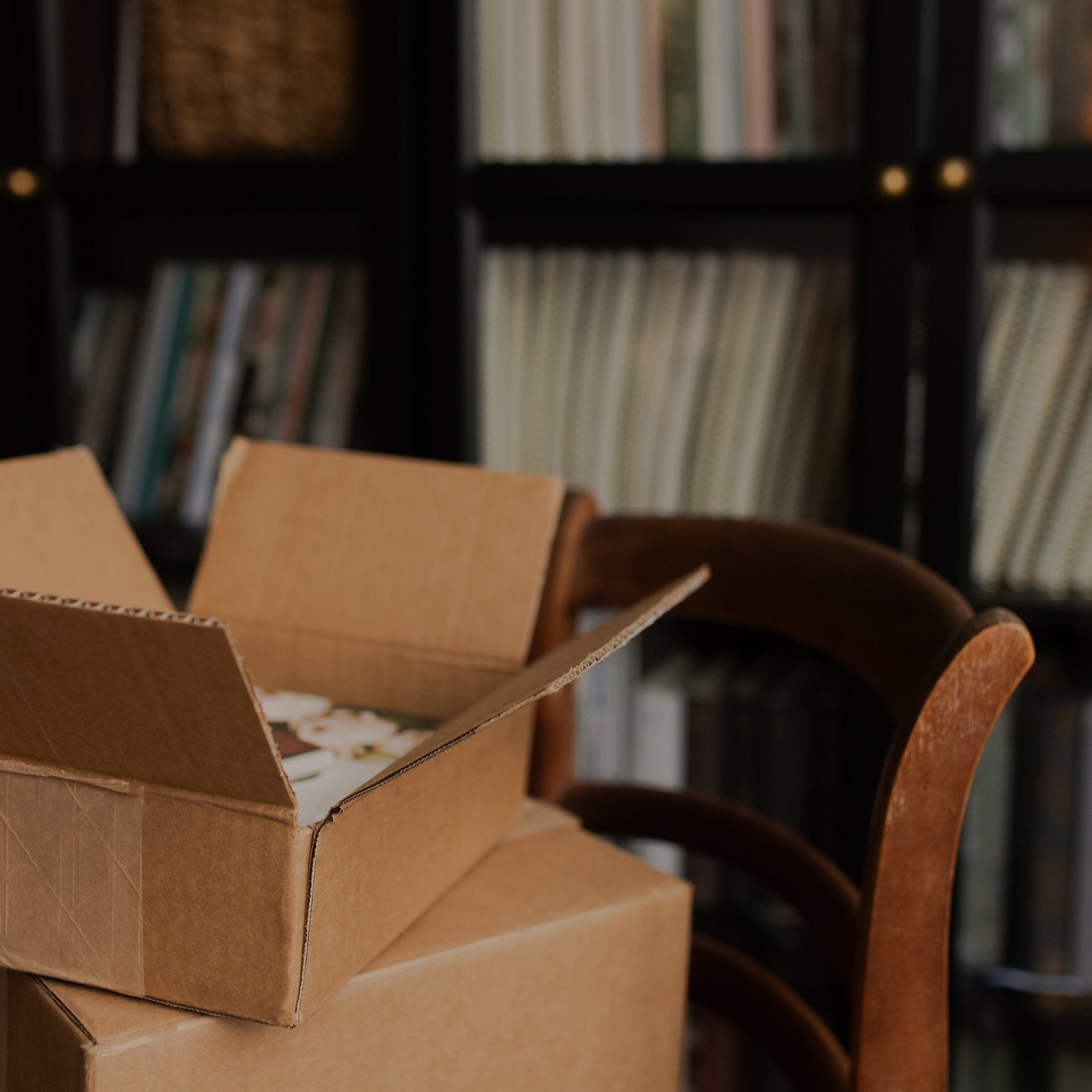 Open cardboard box with items inside, placed on a chair against a blurred bookshelf background.